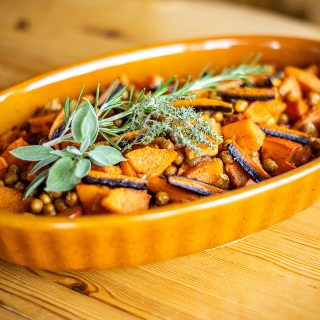 A ceramic baking dish with roasted carrots, sweet potatoes, pumpkin and chick peas, topped with fresh herbs.