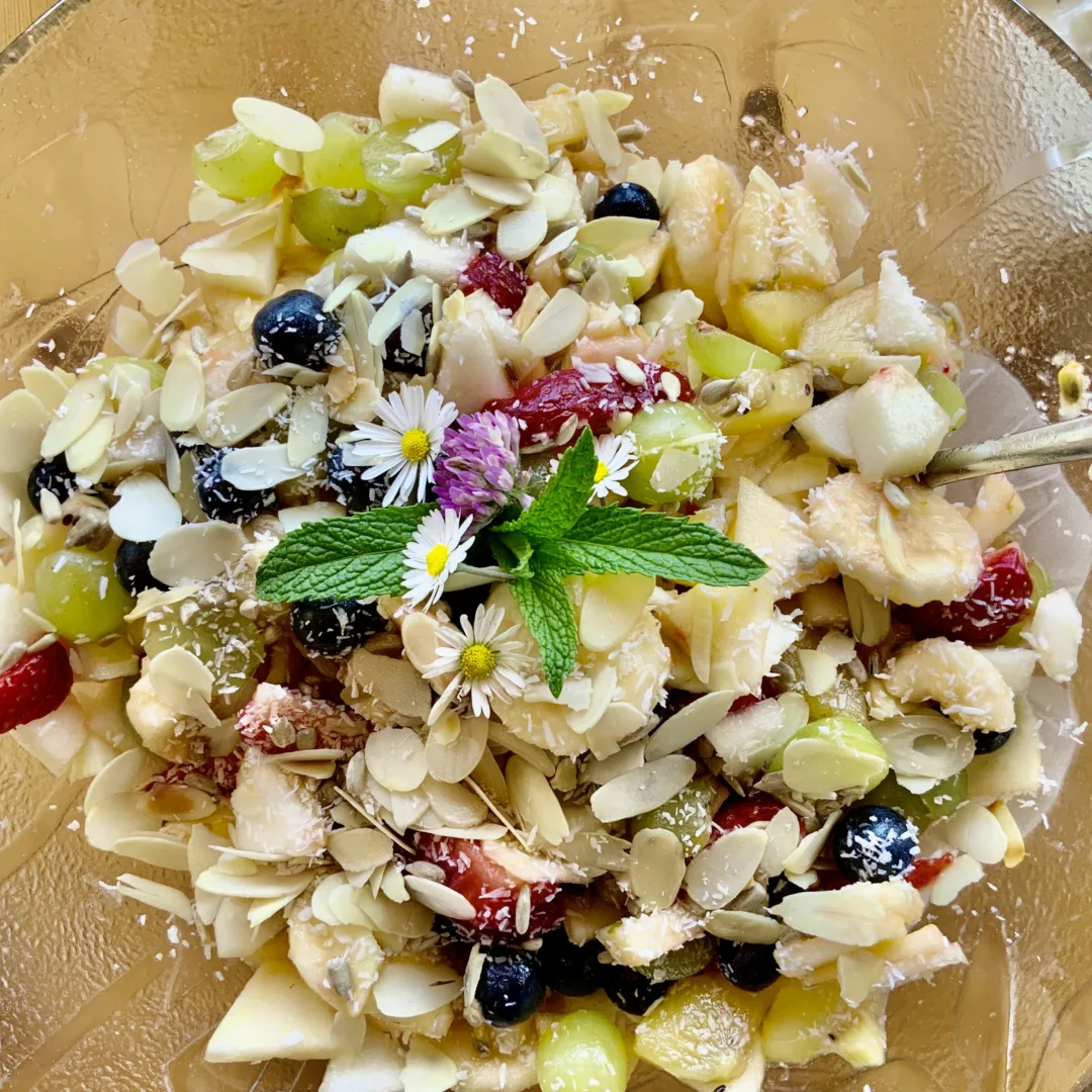 A bowl filled with fresh apple, banana, strawberry, blueberries, and grapes, topped with roasted almond slices and edible flowers.