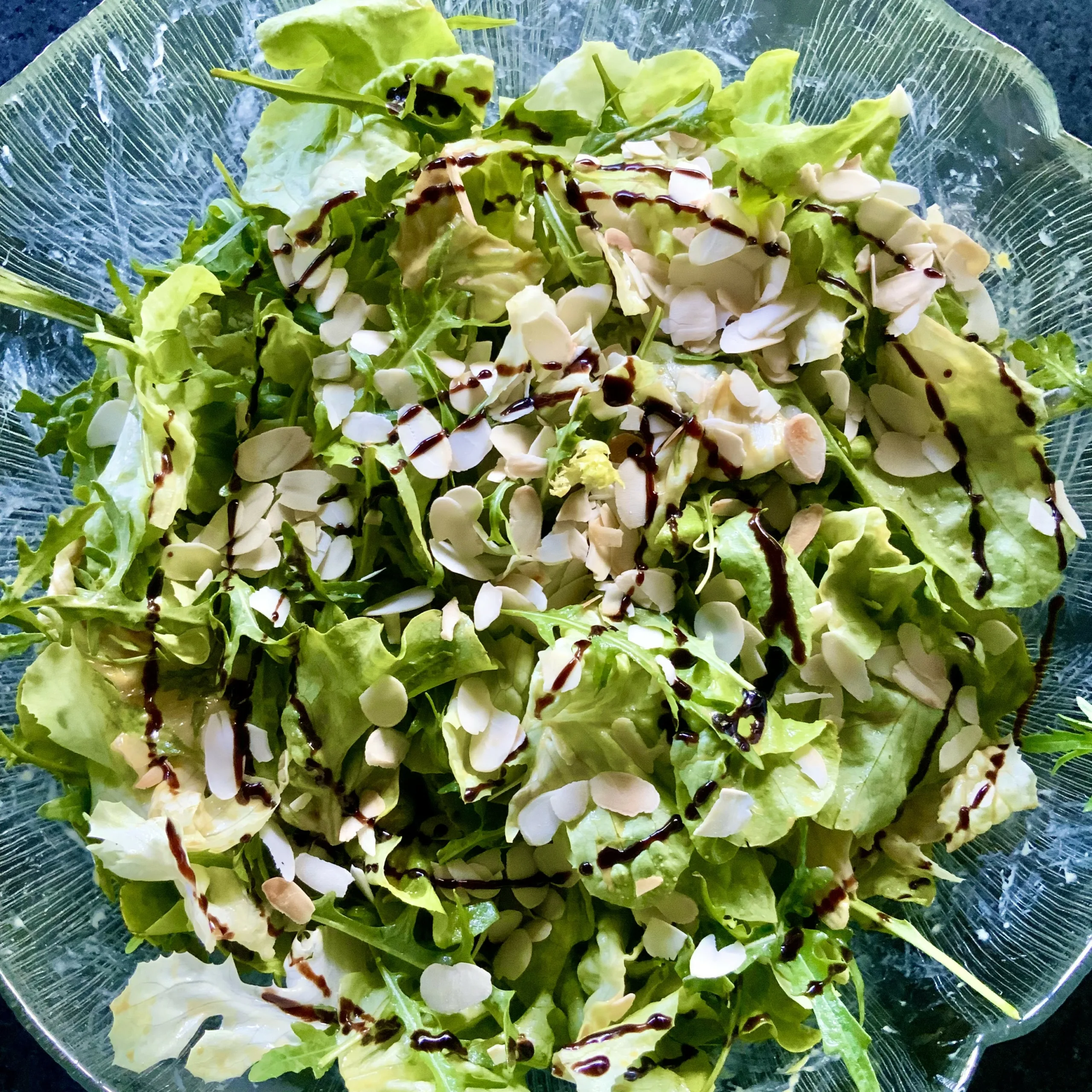 Grüner Salat in einer klaren Glasschüssel, garniert mit gerösteten Mandelblättchen und leicht mit Balsamico-Creme beträufelt.